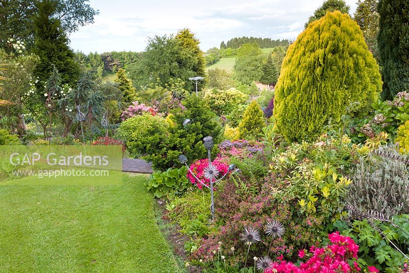 Metal sculptures augment a mixed border at Mount Pleasant Gardens, Kelsall, with the nearby Cheshire hills visible in the distance. Planting includes Azaleas, ferns and Bergenias. June.