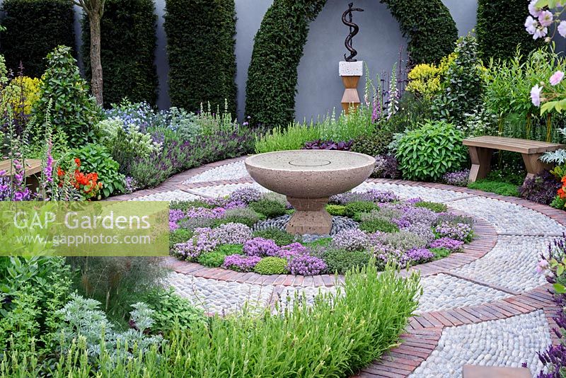 The St John's Hospice Garden, The Modern Apothecary. Extensive planting of herbs around water basin in central circular bed with cobble stone surround. oak seat in border and sculpture of Aesclepius on back wall.  RHS chelsea Flower show 2016