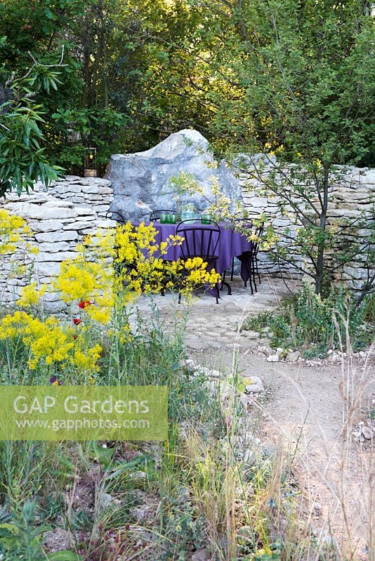L'Occitan Garden, view of earth path leading to a patio with black metal chairs, table with purple table cloth surrounded by Galium verum, Juglans regia. The RHS Chelsea Flower Show 2016, Designer: James Basson MSGD, Sponsor: L'Occitane en Provence