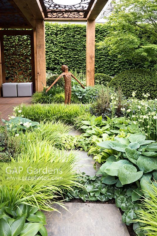 The Morgan Stanley Garden for Great Ormond Street Hospital, path made from paving slabs  leading to patio and bronze sculpture surrounded by Hakonechloa macra 'Alboaurea', Hosta 'Big Daddy', Asarum europaeum, asarabacca, Hosta 'Devon Green',  Taxus baccata ball topiary. RHS Chelsea Flower Show, 2016