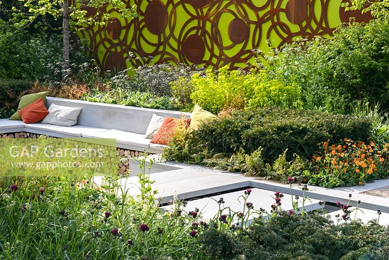 Corylus colurna tree underplanted with Anthriscus sylvestris 'Ravenswing', Dryopertis erythrosora, and Euphorbia robbiae, a bench with log store and colourful cushions, decorative panels by Natasha Webb - The Sunken Retreat, RHS Malvern Spring Festival 2016. Design: Ann Walker, Graduate Gardeners