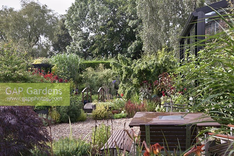 A view across a patio seating area and gravel pathway to a bridge over a small pond surrounded by planting of mixed perennials, shrubs and trees, Cheshire