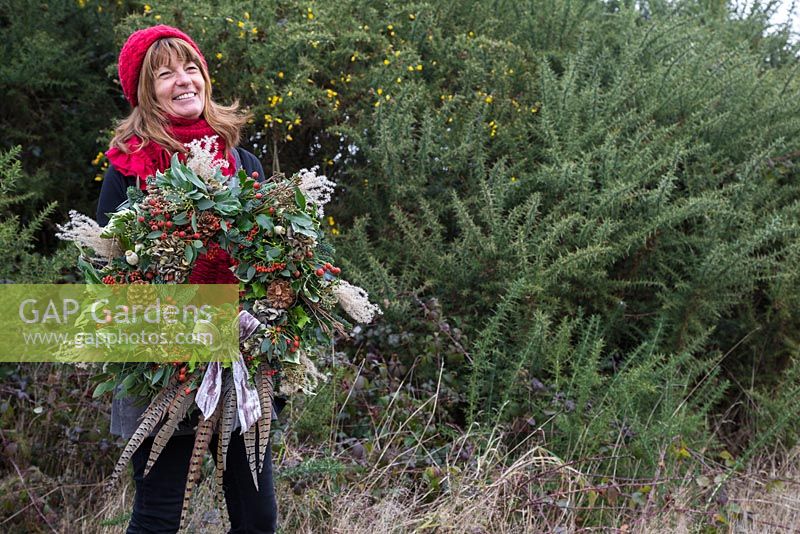 Sheree King holding a traditional christmas wreath made with Eucalyptus gunnii, Rosa 'Bonica' rose hips, Variegated Ivy, Ilex aquifolium, Cotoneaster lacteus, Pinus nobilis, Godetia and Miscanthus sinensis seed heads