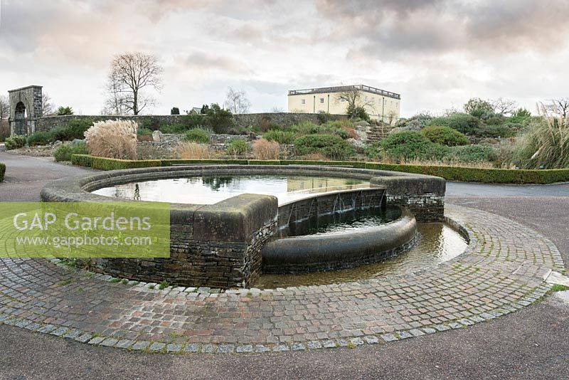 Circular pond framed with stone sets, feeds a curving rill with the Boulder Garden and Principality House behind in December
