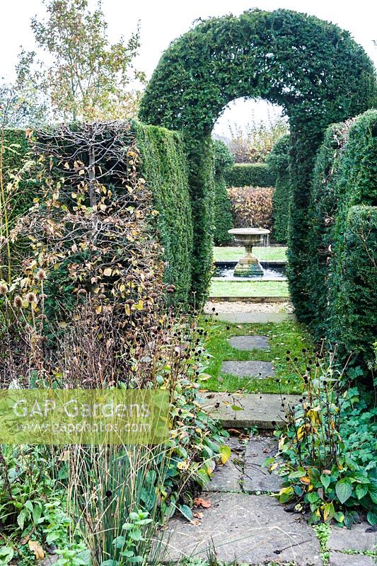 A clipped yew arch frames a view through the garden between yew and hornbeam hedges to an urn, overflowing with water, sitting in a pond.