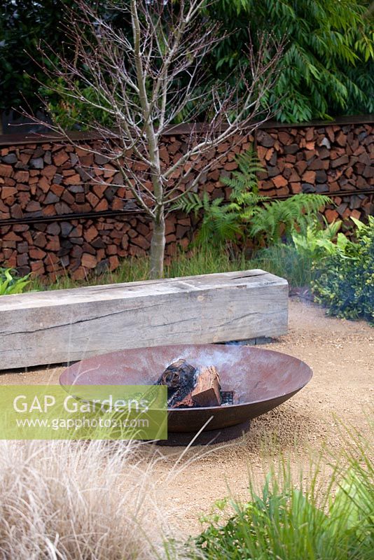 A fire pit with on a paved area in front of a timber garden bench surrounded by a planting of mixed plants and grasses