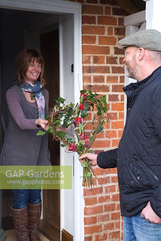 A man handing a rustic Valentine's wreath to his partner. Made with Hedera helix, Willow stems, Birch twigs, Cornus sanguinea 'Midwinter Fire', Tulip 'Rococo', Viburnum tinus and Chamelaucium