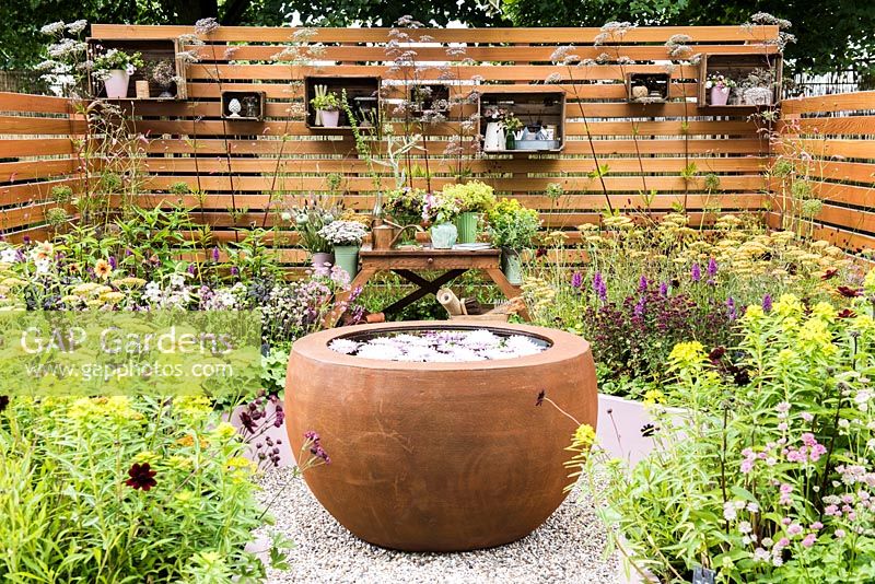 View of the garden with circular water feature, wooden work table with bouquets in pots and containers surrounded by wooden fence and summer flowers including Achillea 'Terracotta', Alchemilla mollis, Astrantia 'Pink Pride', Euphorbia schillingii, Valeriana officinalis. Katie's Lymphoedema Fund: Katie's Garden, Designers: Carolyn Dunster, Noemi Mercurelli, RHS Hampton Court Palace Flower Show 2016 