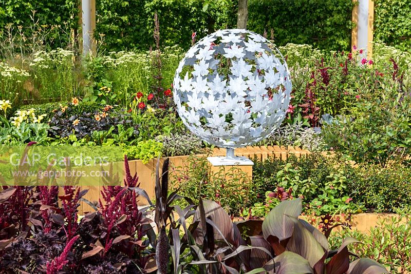 A round sculpture with leaf pattern by Paul Richardson with planting of Amaranthus 'Red Army' and purple-leaved cannas. Witan Investment Trust Global Growth Garden, RHS Hampton Court Flower Show 2016