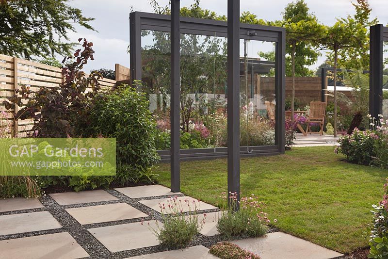 A patio with gravel between stone slabs, lawned area, framed glass panels and a dining area beyond - Through The Looking Glass at RHS Tatton Park Flower Show 2016