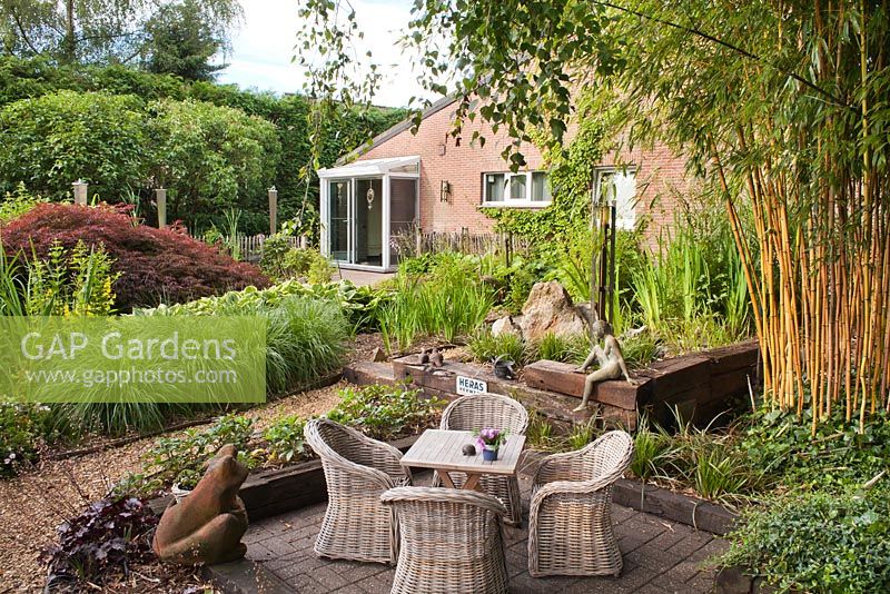 Patio with garden furniture. Planting includes bamboo Phyllostachys vivax 'Aureocaulis', Vinva periwinkle, Heuchera 'Blackberry Jam', Lysimachia punctata, Pennisetum alopecuroides 'Hameln', Iris sibirica. Sempre Verde Tuin in Belgium.