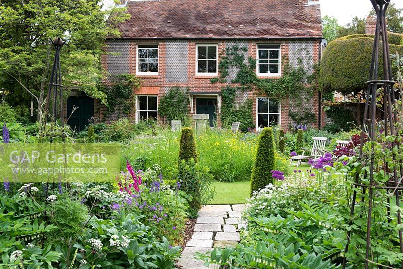 A 19th century cottage with circular lawn, raised wildflower mound, yew topiary and mixed borders with Anthriscus 'Ravenswing', Allium 'Purple Sensation', Geranium sylvaticum 'Album' and Gladiolus byzantinus.