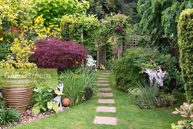 A stepping stone path leads over lawn, past bench, and beneath clematis arch to work area. On left, bubbling urn water feature and purple acer.
