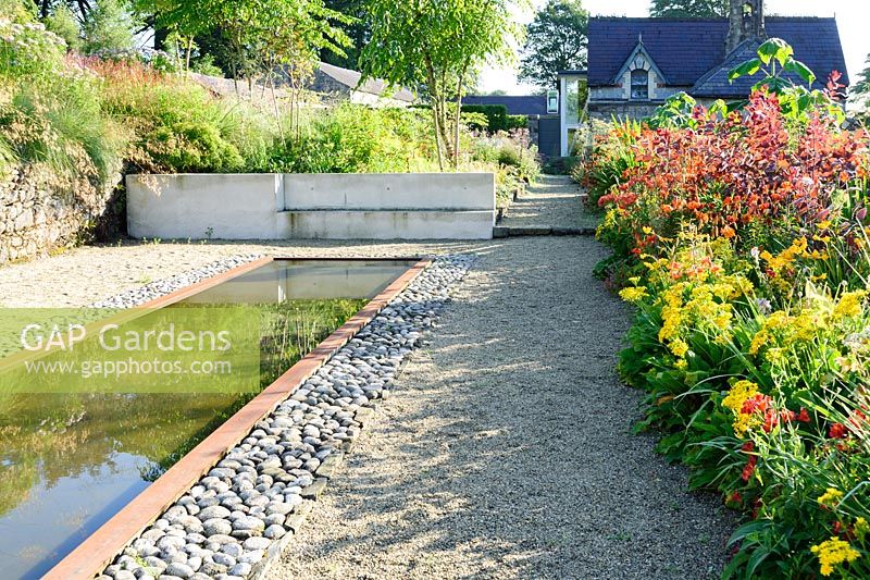 Reflecting pool edged with rusted steel and inset pebbles beside hot beds full of vivid red, orange and yellow herbaceous perennials and shrubs including yellow Leontodon rigens, orange Lilium lancifolium, Cotinus 'Grace', Dahlia australis and Crocosmia 'Lucifer'.