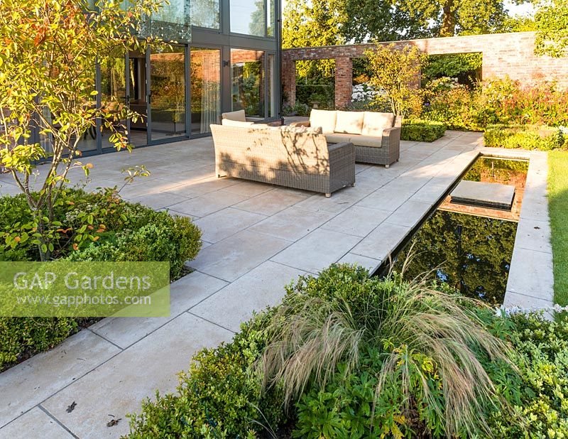 A patio area, with seating and a rill, in a modern Cheshire country garden, designed by Louise Harrison-Holland. Planting includes Amelanchier lamarckii, box hedging and Stipa tenuissima