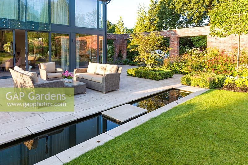 A patio area, with seating and a rill, in a modern Cheshire country garden, designed by Louise Harrison-Holland. Planting includes box hedging, Amelanchier lamarckii and Stipa tenuissima