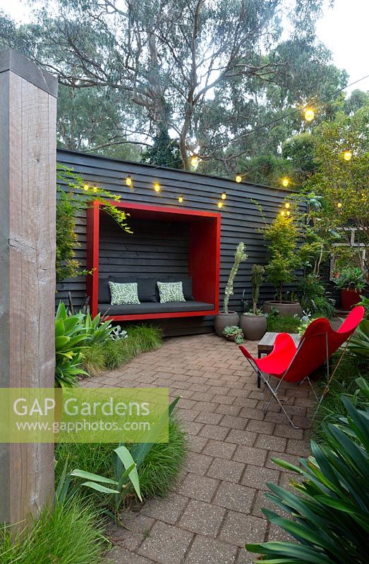 Paved area featuring a black painted timber wall with a red sitting pod attached to it a red canvas retro butterfly chair and a collection of large pots with a maple and various succulents.