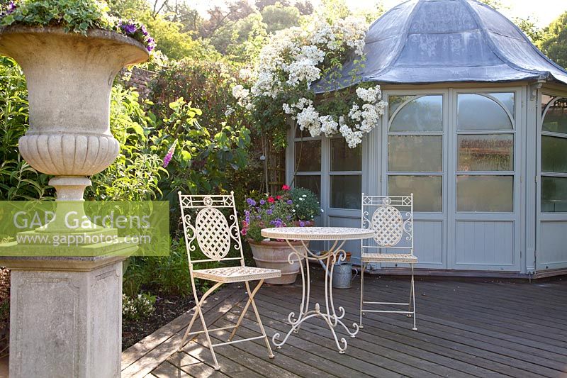 Decorative metal furniture on decked patio with stone urn and summerhouse with Rosa