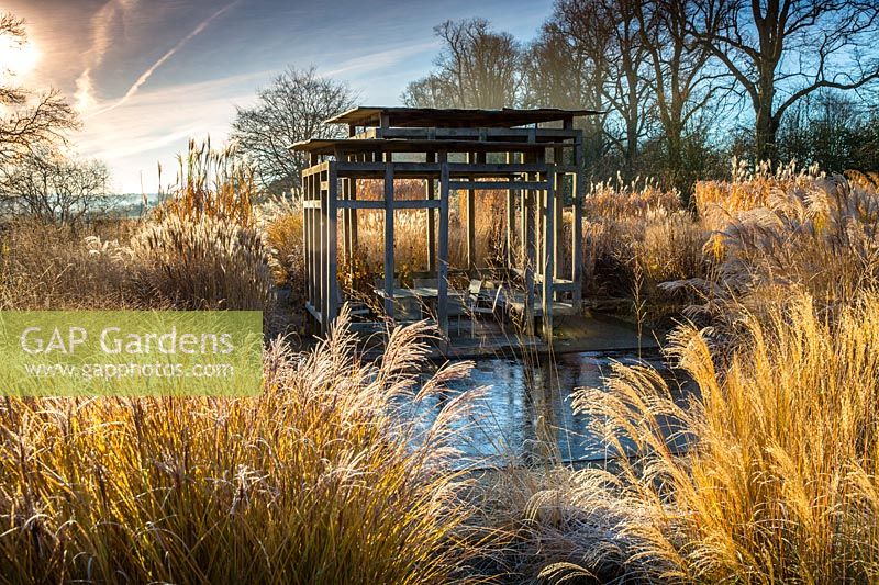 View across reflective pool and swathes of Miscanthus and other tall grasses to seating area within modern oak summerhouse in the minimalist front garden in Winter at Bury Court Gardens, Hampshire. Designed by Christopher Bradley-Hole.