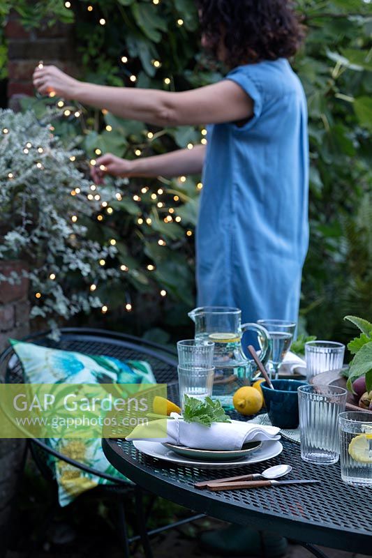 Dusk falls on the outdoor dining area under a fig tree. Woman puts fairy lights in the trees to create a cosy setting and light up the area. Table laid with greens and fresh whites and napkins dressed with a geranium leaf
