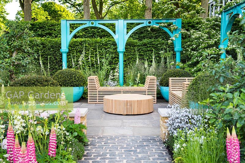 View of blue arches, seating area with wooden benches, chairs, table and huge blue pots with Taxus baccata topiary spheres surrounded by Lupinus 'Blossom' and Lupinus 'Rachel de Thame',  The Sir Simon Milton Foundation Garden: '500 years of Covent Garden' - RHS Chelsea Flower Show 2017 - Designer: Lee Bestall - Sponsor:  Capco Covent Garden