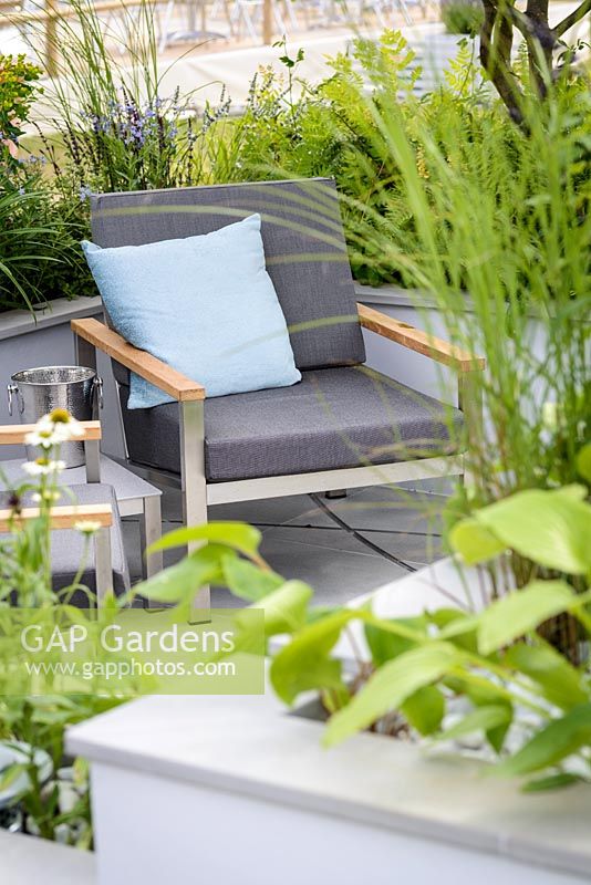 Contemporary  sunken patio with seating area surrounded by raised beds with grey armchairs and blue cushions - The Urban Rain garden.  RHS Hampton Court Palace Flower Show 2017. Design: Rhiannon Williams. Sponsors: Landform