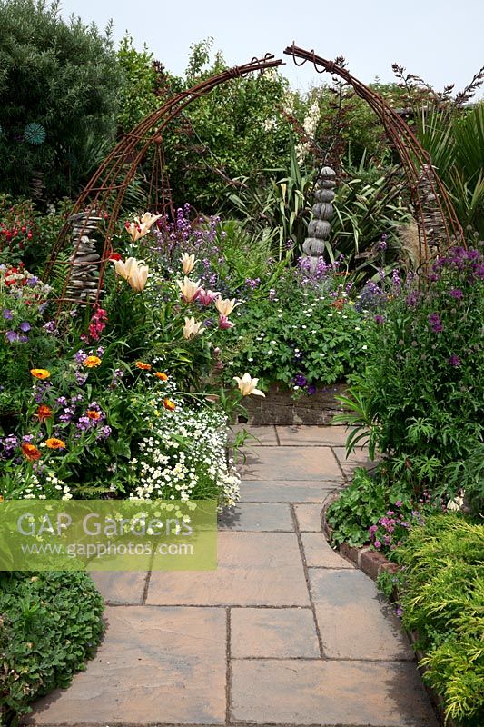 Pathway and view to rusty metal arch  with Calendula - Marigolds, Centaurea montana 'Purpereum', Petunias, Lilies, Anthemis,Fuschia 'Empress of Prussia', Campanula, Erysimum 'Bowle's Mauve',   and backdrop of Phormium with ball sculpture - Driftwood