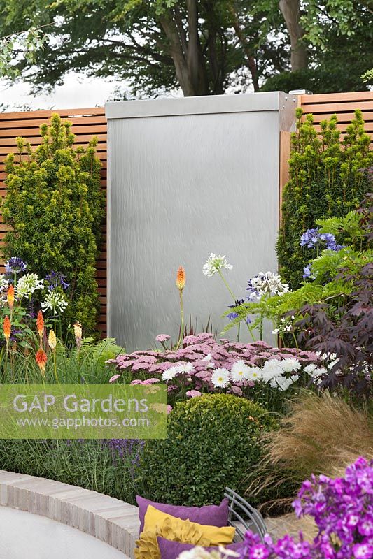 A stainless steel sheet water wall cascade flanked by Taxus baccata 'Fastigiata Aureomarginata' and  Kniphofia 'Royal Standard', Achillea 'Lilac beauty' and Leucanthemum x superbum 'Aglaia' in front in the 'Facing Fear, Finding Hope' Garden at Tatton RHS Flower Show 2017