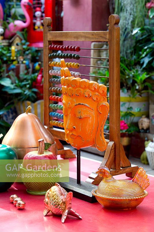 A collection of eclectic brightly coloured ornaments on a red retro laminex table underneath a covered outdoor entertaining area featuring metal fruit, an childs abacus and an Thai style head ornament painted orange.