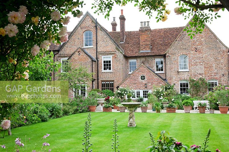 View of the house through rose covered pergola with dolphin stone birdbath on lawn and pot filled terrace.