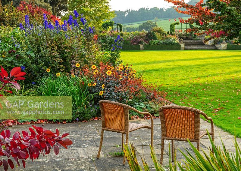 Patio with wicker seats and border with  Dahlia 'Moonfire', Aconitums - Bourton House Garden, Gloucestershire

