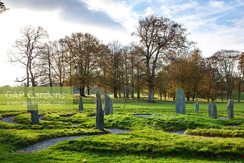 Stone circle comprising a winding labyrinth with monoliths of slate set in grass, backdrop of deciduous trees in autumn
