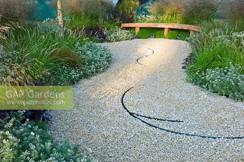 Gravel path leading to a wooden bench seat, edged with borders planted with grasses and white flowers