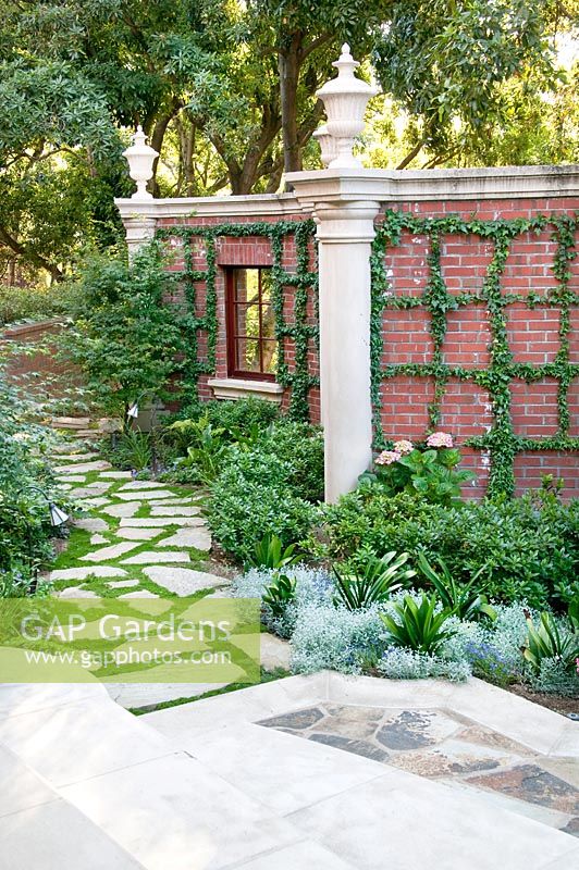 Stone walkway with shrubs and perennials, wall with terra cotta tiles and Hedera, columns and urns