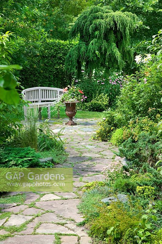 Garden path with shrubs and perennials, garden bench and planted iron urn