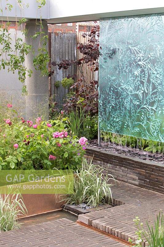 The Silent Pool Gin Garden, view of sunken garden with glass wall water feature, corrugated metal fencing, recycled concrete pillars, clay brick wall and paving, roses, grasses on  copper raised bed – Designer: David Neale - Sponsor: Silent Pool Gin