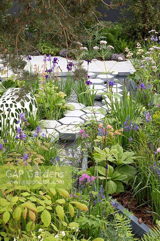 The Manchester Garden, view of sculptures and cottage garden planting, with hexagonal stepping stones over the pond, bordered by Rodgersia, Iris sibirica 'Persimmon, Iris sibirica 'Caeser's Brother', and Iris sibirica 'Harpswell Happiness' - RHS Chelsea Flower Show 2019 - Designer: Exterior Architecture, exteriorarchitecture.com 