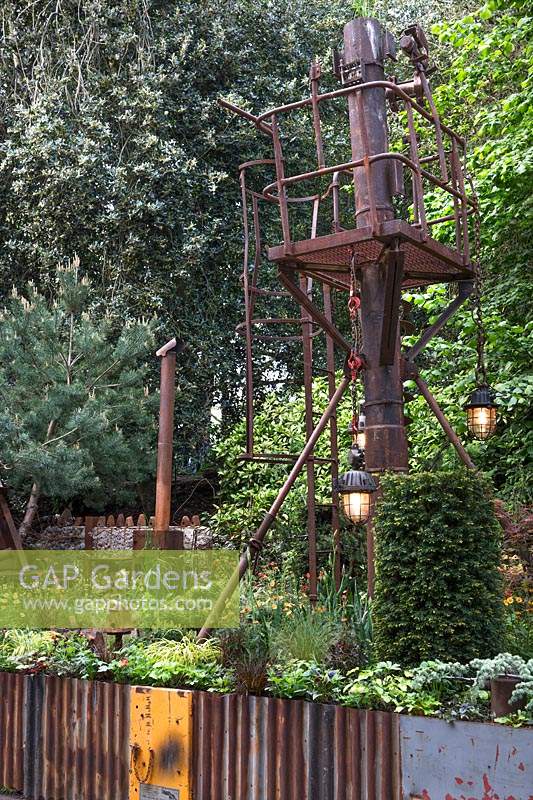 Walker's Forgotten Quarry Garden. Rusted abandoned industrial tower with plants including Taxus baccata - Yew topiary pillars and orange Geum. Design: Graham Bodle. Sponsor: Walker's Nurseries