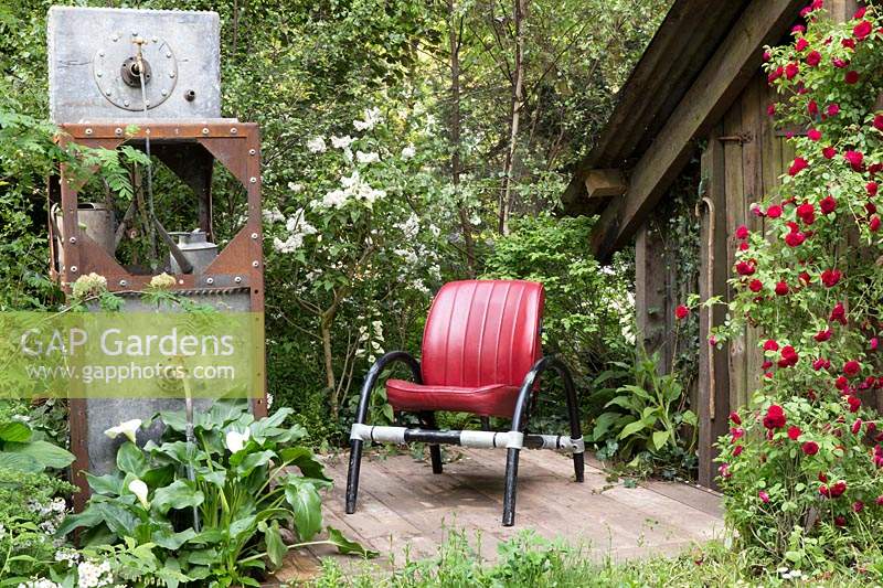 The High Maintenance Garden for Motor Neurone Disease Association Garden, view of red garden chair seat on small patio, climbing red rose Rosa 'Chevy Chase', galvanised metal container with old tap water feature under planted with Zantedeschia aethiopica, Arum Lily, Calla Lily. Design: Sue Hayward - Sponsor: Martin Anderson 