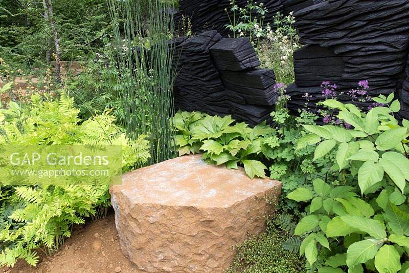 The M and G Garden, view of green planted woodland garden with shade loving plants English ironstone stone seat, charred oak walls sculpture by Johnny Woodford – Designer: Andy Sturgeon - Sponsor: M and G 