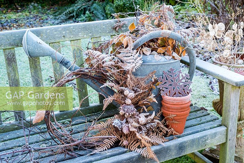 Frosty watering can with natural wreath of dried seedheads, beech leaves and ferns on garden bench