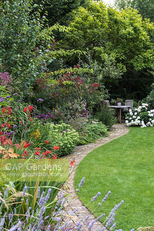 A curving brick path between a mixed border and lawn, leads to seating area
