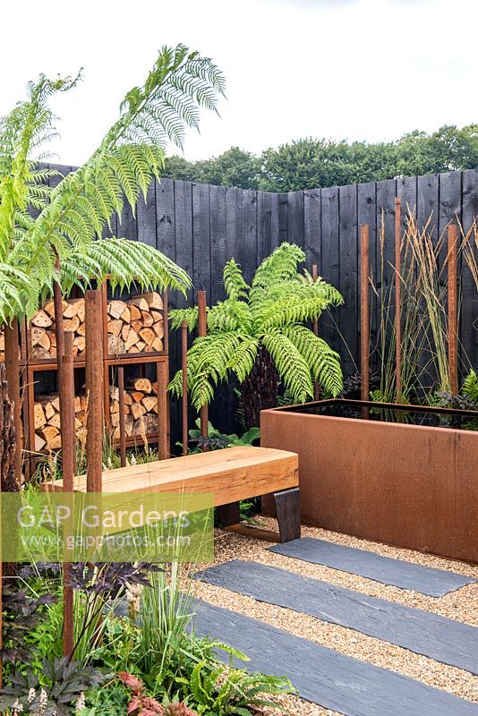Slate paving with gravel leading to an oak bench, Corten water container, Dicksonia antarctica and log store surrounded by a burnt effect fence - Bee's Gardens, The Penumbra, RHS Tatton Park Flower Show 2018