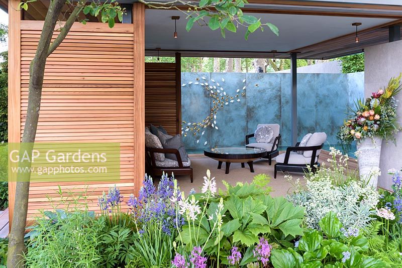 Armchairs and table in timber pavilion with eddy art wall and planting of Dodecatheon meadia, Euphorbia characias subsp. wulfenii 'White Swan' and Camassia leichtlinii 'Maybelle' - The Morgan Stanley Garden for the NSPCC - Sponsor: Morgan Stanley - RHS Chelsea Flower Show 2018