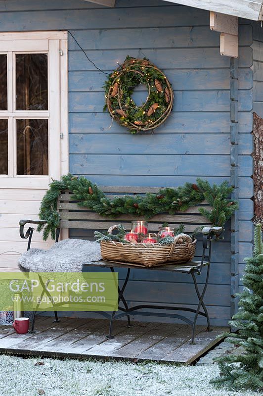 A snowy bench in front of a blue summer house decorated with a fir garland, a basket and a wreath on the wall 

