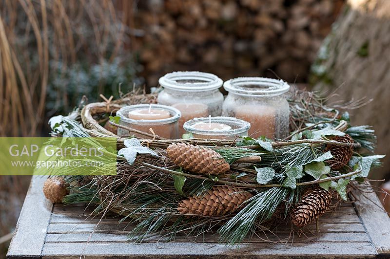 An Advent wreath on a table in the garden bonded out of Pinus strobus cones, 
pine green, ivy Hedera helix, clematis cirrus, covered by powder snow or white 
frost. Inside the wreath you find 4 candles in lanterns.