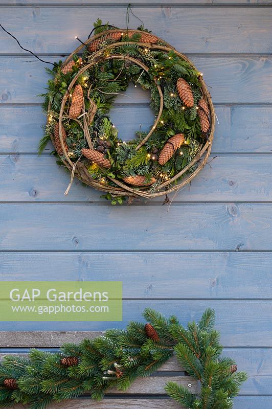 A wreath hanging on the wall of blue summer house featuring fairy lights, 
mixed greenery, and pine cones 