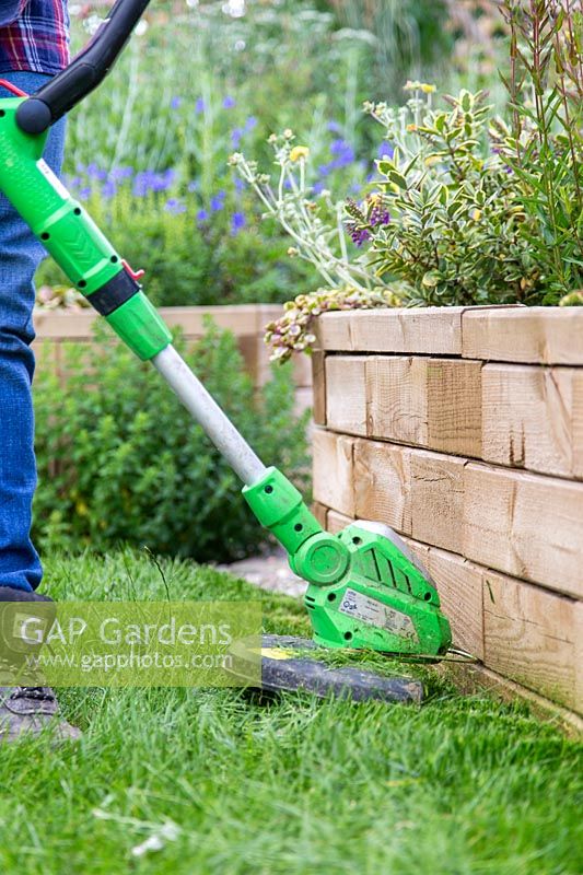 Woman using strimmer to cut long grass along edge of raised bed