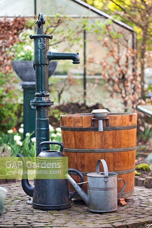 View of vintage watering pump in the garden with tap, barrel and watering cans.
