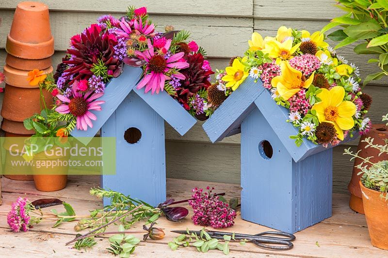 Pair of wooden birdboxes with flower embellished roofs on a wooden bench with cut flower heads and scissors
